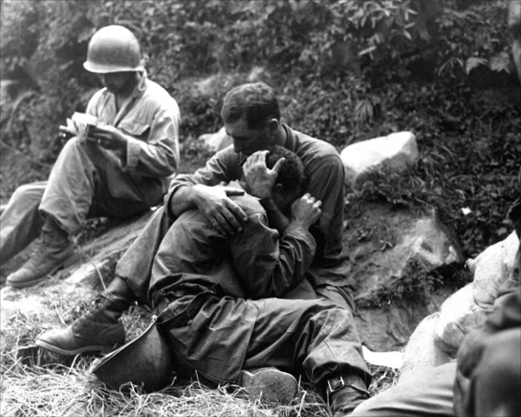A grief stricken American infantryman whose buddy has been killed in action is comforted by another soldier. In the background a corpsman methodically fills out casualty tags, Haktong-ni area, Korea. August 28, 1950.