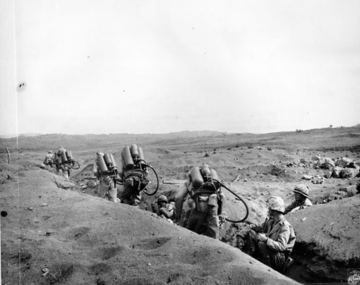 Riflemen lead the way as flame-throwing Marines of the Fifth Division, crouched with the weight of their weapons, move up to work on a concentration of Japanese pillboxes. Photo: USMC
