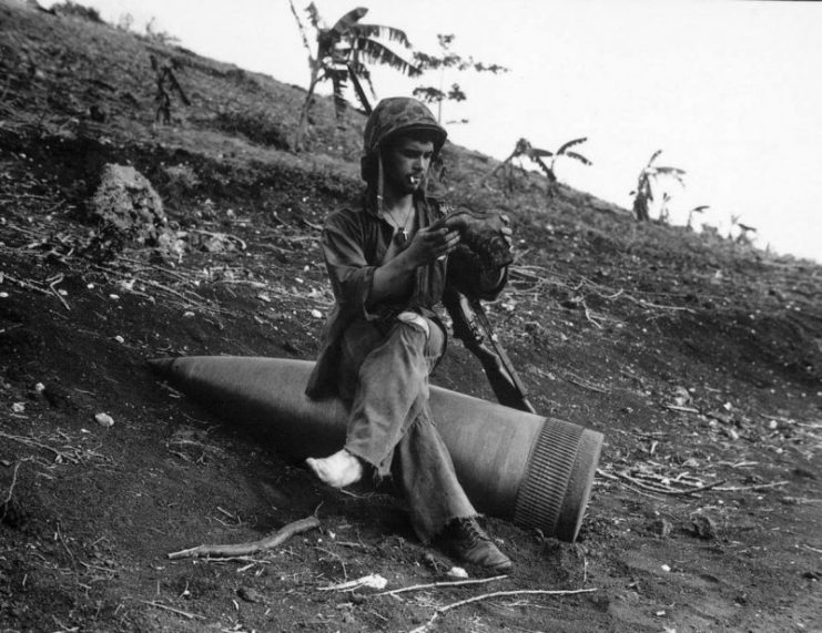 A U.S. Marine sits on an unexploded shell of a 16″/50 caliber Mark 2 gun to inspect a hole in the sole of his boot during the Battle of Saipan.