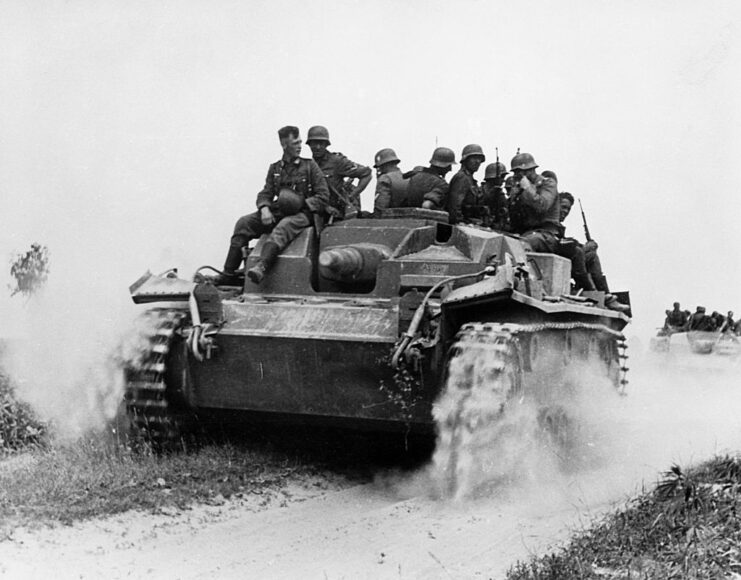 German soldiers riding on a tank that's driving along a dirt road