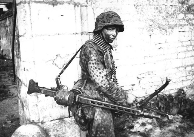 A German Waffen SS soldier involved in heavy fighting in and around the French town of Caen in mid-1944. He is carrying an MG 42 configured as a light support weapon with a folding bipod and detachable 50-round belt drum container.Bundesarchiv – CC-BY SA 3.0