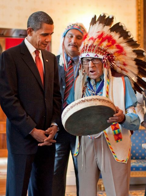 Joe Medicine Crow in full feathered headdress plays a drum for U.S. President Barack Obama.