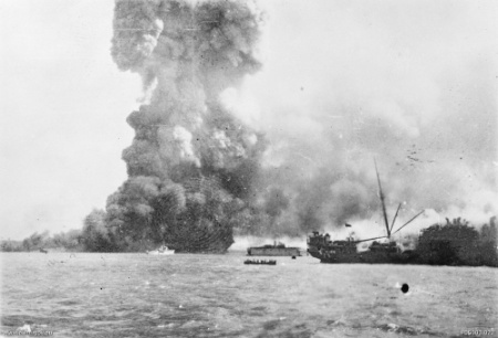 MV Neptuna explodes at Stokes Hill Wharf. In front of the explosion is HMAS Vigilant which is undertaking rescue work. In the centre background is the floating dry dock holding the corvette HMAS Katoomba. In the foreground is the damaged SS Zealandia.