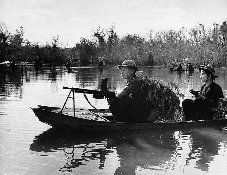 Viet Cong Guerrillas, led by North Vietnamese Regulars, bear automatic weapons and use leafy camouflage as they patrol a portion of the Saigon River in small boats somewhere in South Vietnam