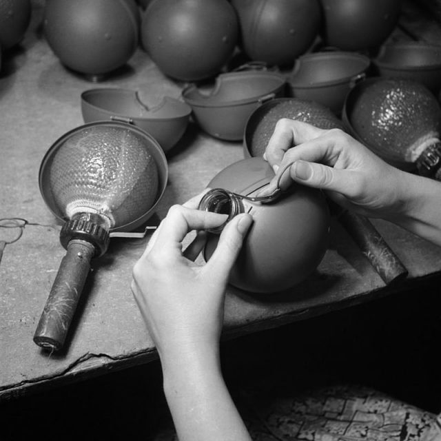 A close-up view showing the protective casing being clipped into place around the glass flask of a sticky bomb at a workshop, somewhere in Britain. The casing is held in place by a narrow metal band around the neck of the flask. Once this clip is released, the two halves of the casing fall away, exposing the adhesive surface of the bomb.