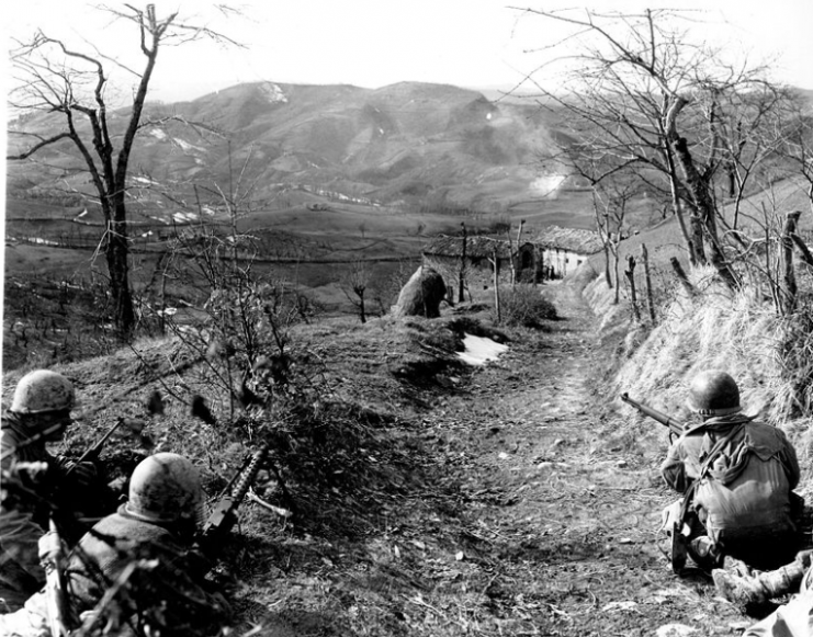 A machine gunner and two riflemen of Co K, 87th Mountain Infantry, 10th Mountain Division, cover an assault squad routing Germans out of a building in the background. Sassomolare Area, Italy. Porretta Moderna Highway. 4 March 1945.