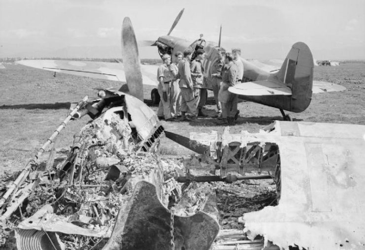 A conference of ground crews and pilots of No. 111 Squadron RAF by one of their Supermarine Spitfire Mark VCs, seen over the wreckage of an Italian fighter, destroyed in its blast bay on the edge of the airfield