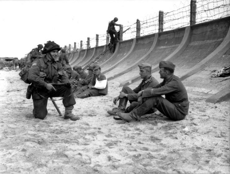Corporal Victor Deblois of the Chaudière Regiment questions squatting two German prisoners captured by Canadian troops at Juno Beach on D Day. Other prisoners are also sitting along the anti-tank wall.