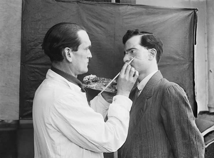 The Development of Reconstructive Plastic Surgery during the First World War. Captain Francis Derwent Wood holds an artist’s palette as he adds the finishing touches to a patient’s new facial plate. He is trying to make the mask blend in with the patient’s skin to make it look as life-like as possible, 3rd London General Hospital.
