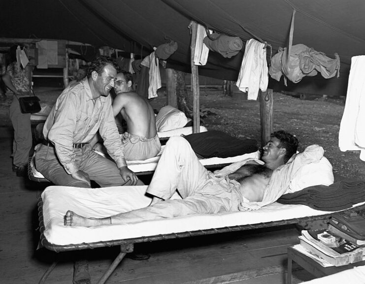 John Wayne sitting on a cot while speaking with a serviceman lying down
