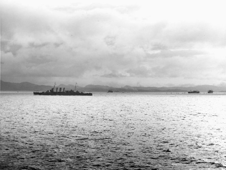 HMAS Canberra (center left) protects three Allied transport ships (background and center right) unloading troops and supplies at Tulagi.