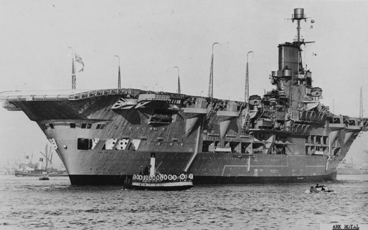 Ark Royal’s flight deck overhangs the stern. Her unusual height above the waterline is visible in comparison with the tugboat.