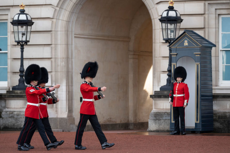 Three Grenadier Guards walking past an entryway at Buckingham Palace while another stands watch