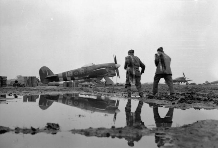 Two armourers of No 440 Squadron, Royal Canadian Air Force, trudge through the mud of an airfield to re-arm a Hawker Typhoon fighter-bomber.
