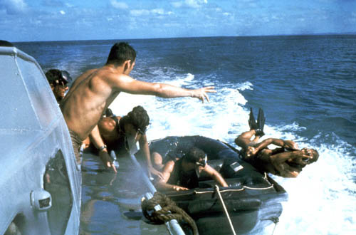 A US Navy Sea-Air-Land (SEAL) team member enters the water from an inflatable boat while preparing for a stringline recovery operation. The raft is being towed by another boat.