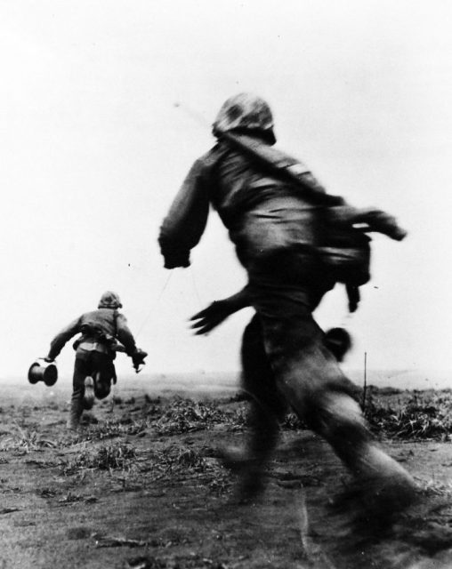 On Iwo Jima, two Marine wiremen of the Fifth Division race across an open field, under fire, to establish field telephone contact with the front lines.