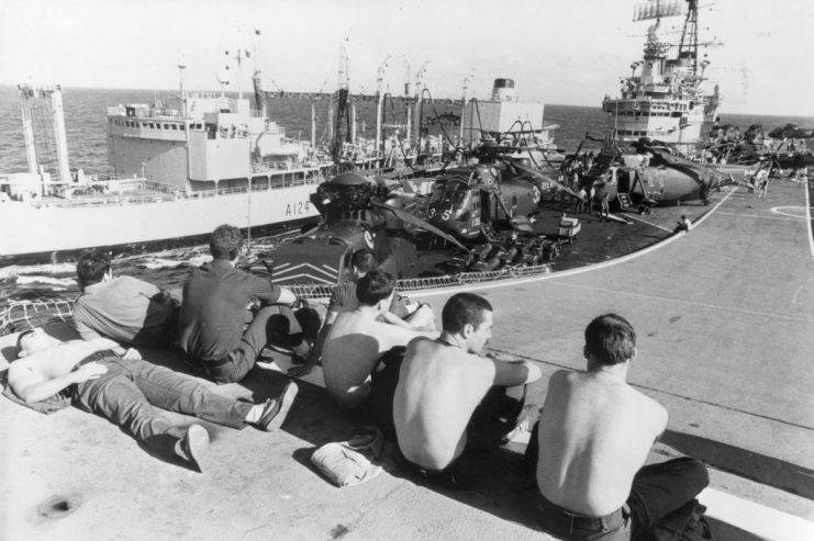 19th April 1982: Marines relaxing on the ‘ski jump’ ramp of HMS Hermes, as she is replenished at sea by a Royal Fleet auxiliary vessel, on the way to the Falkland Islands, during the Falklands War. Westland Sea King helicopters can be seen on deck. (Photo by Fox Photos/Getty Images)