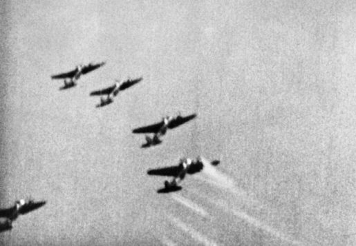 A still from camera-gun film taken from a Supermarine Spitfire Mark I of No. 609 Squadron RAF, flown by by Pilot Officer J D Bisdee, as he dives on a formation of Heinkel He 111s of KG 55 which have just bombed the Supermarine aircraft works at Woolston, Southampton. The rearmost aircraft of the leading ‘staffel’ receives a burst of machine gun fire from Bisdee, as shown by the streaks of light from the tracer bullets. Its port engine is also on fire. [© IWM (CH 1827)]