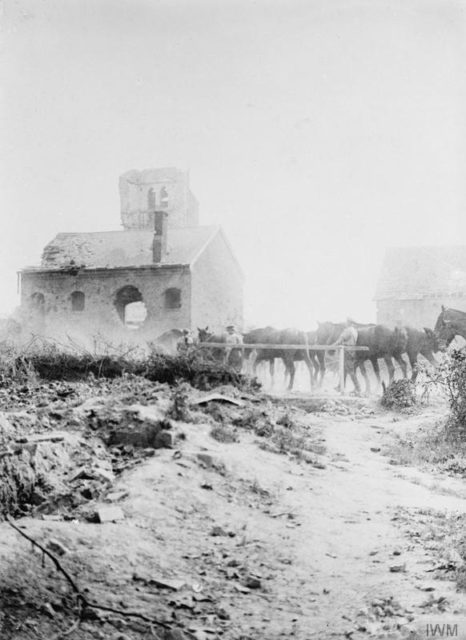A house with a shell hole right through it by a ruined church with an observation balloon visible high in the background. Horses are being watered in the foreground.