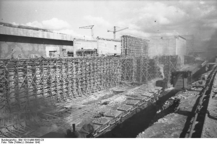 A new harbor basin is being excavated in front of the submarine boxes. Small tilting lorries move the earth away.