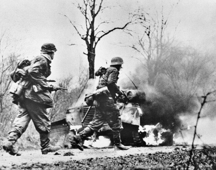 Two German soldiers walking by a burning tank during the Battle of the Bulge