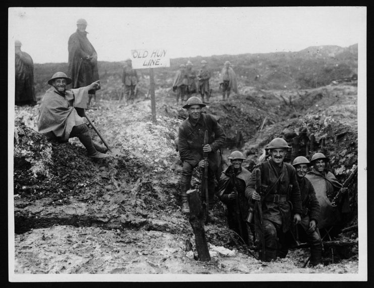 British soldiers at the old German Front Line, during World War I. In front of a mound and standing in a network of trenches are groups of soldiers, mostly smiling and laughing.