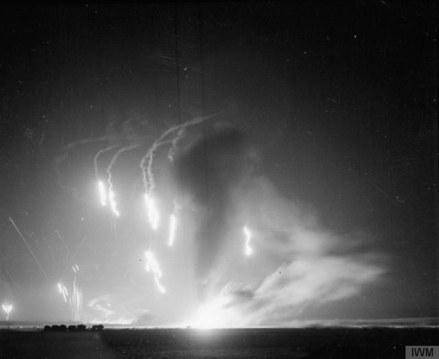Chandelier flares light up an Allied airfield during a night raid by Axis bombers. Bombs are bursting and a column of smoke rises into the night sky from a fire. [© IWM (CNA 1293)]