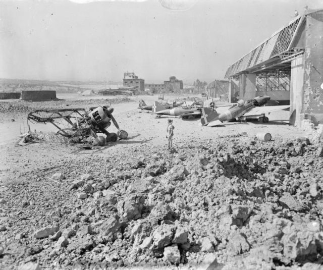 Wrecked and damaged Italian fighters outside bomb-shattered hangars at Catania, Sicily, under the scrutiny of an airman, shortly after the occupation of the airfield by the RAF. [© IWM (CNA 1352)]