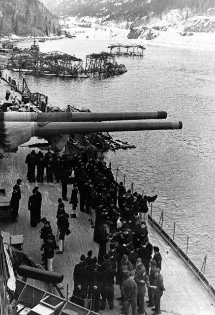 Crewmen on board the battleship, while she was moored in a Norwegian fjord, circa 1942-44. One of the ship’s 380mm (15 in) gun turrets is trained abeam