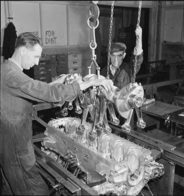 Two workers at a Merlin aircraft engine factory gently lower a Crankshaft Assembly into the Crank Case.