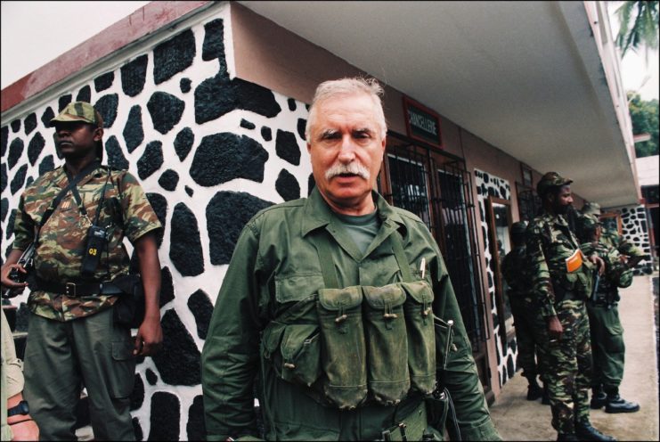 Bob Denard standing outside of a building flanked by guards