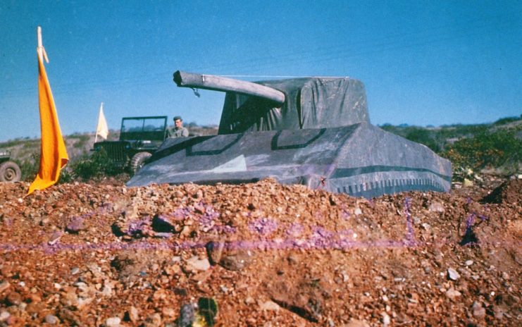 Inflatable dummy tank positioned in a mound of dirt, with a soldier sitting nearby in a Jeep