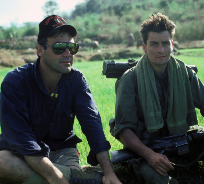 Charlie Sheen and Oliver Stone sitting together on the grass