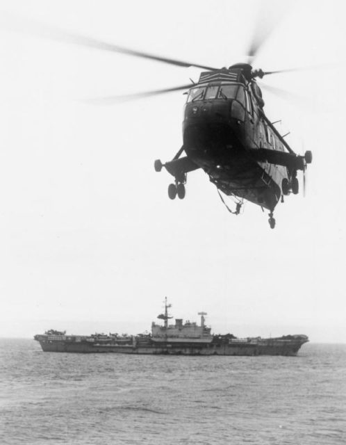A sea king helicopter flying away from the HMS Hermes