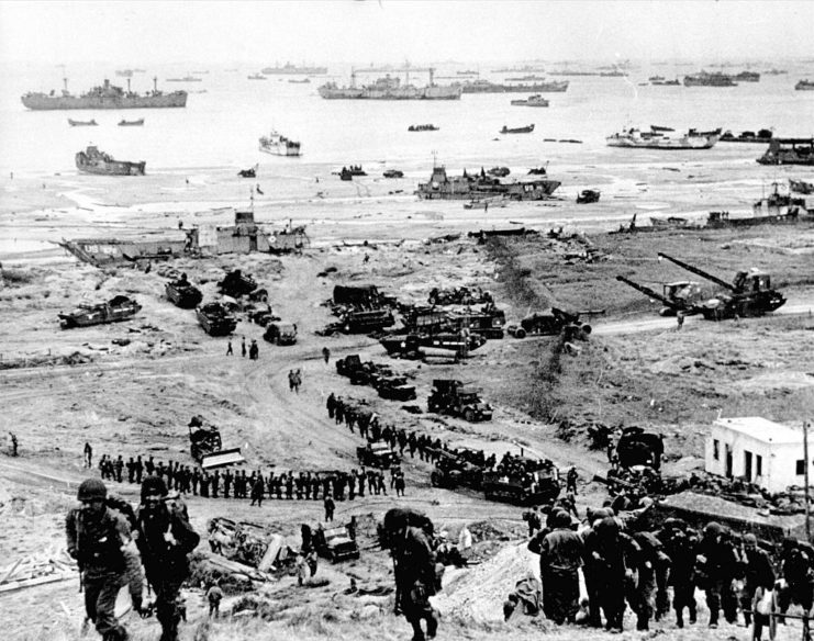 Allied troops walking along Omaha Beach while boats sit in the water