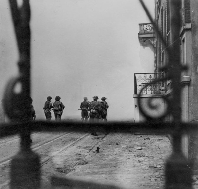 Five British soldiers walking down a street shrouded in dust and smoke