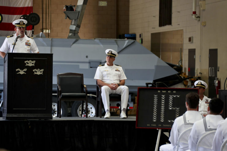 Commander Brad Geary standing behind a podium with a sailor sitting behind him