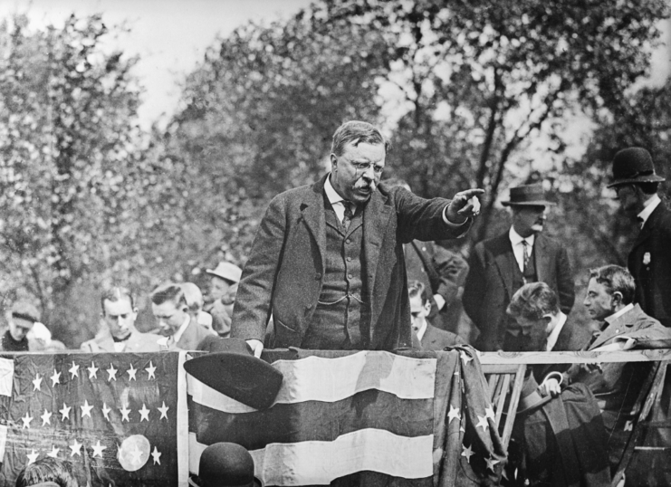 Theodore "Teddy" Roosevelt standing on a podium, with men sitting around him