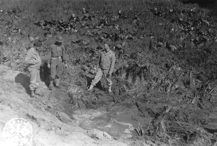 Three US servicemen standing around a shell crater