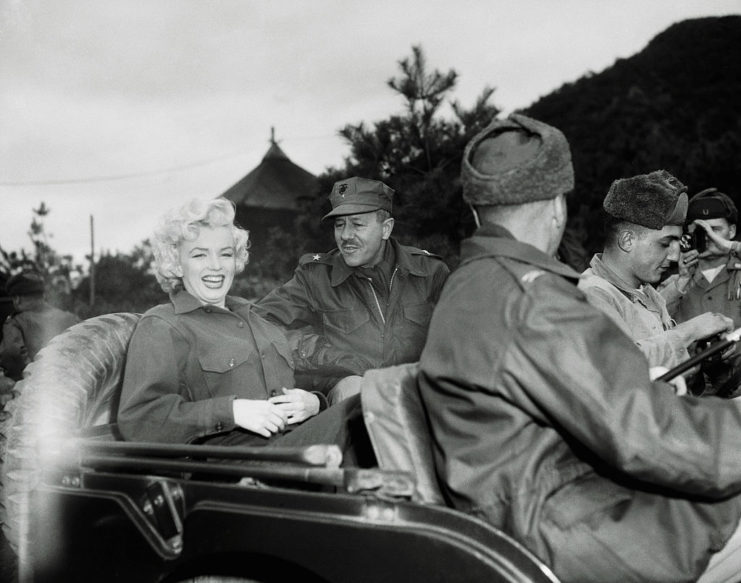 Marilyn Monroe riding in a Jeep with Brigadier General Robert Hobaboom and other GIs