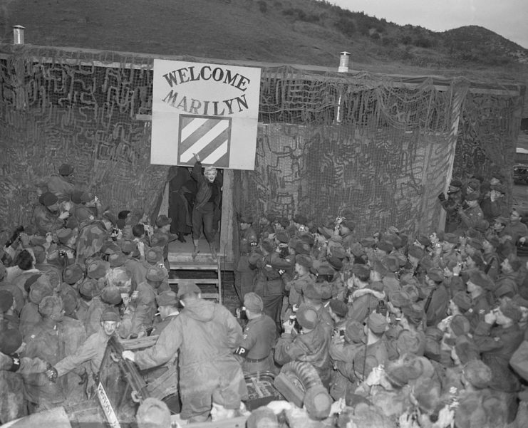 Marilyn Monroe waving at a crowd of troops who are waiting for her by a doorway