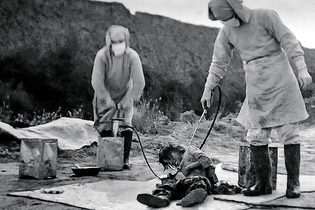 Two people in protective gear spraying a substance on another person