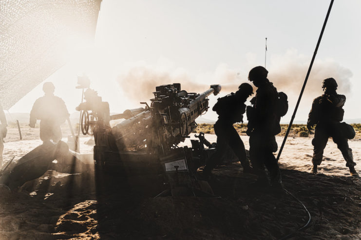 Four Marines standing in shadow beside a M777 Howitzer