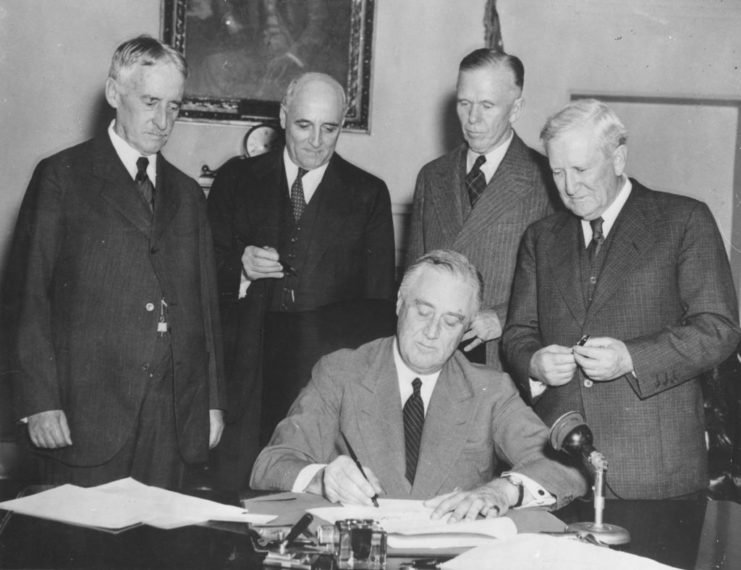 Henry Stimson, Andrew May, George C. Marshall and Morris Sheppard standing above President Franklin Roosevelt as he sits at a desk