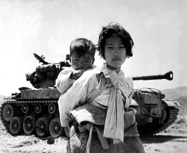 Korean War refugee with her baby, standing in front of a tank