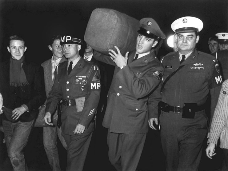 Elvis Presley walking with a group of people while carrying his rucksack over his shoulder