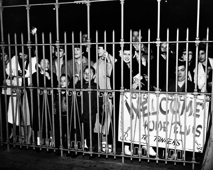 Fans waiting behind a fence, with one holding a sign reading, "WELCOME HOME ELVIS"
