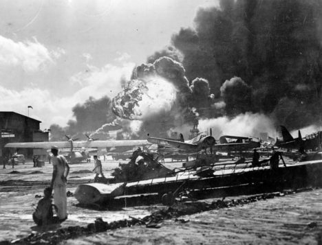Sailors standing among airplanes while smoke billows in the distance