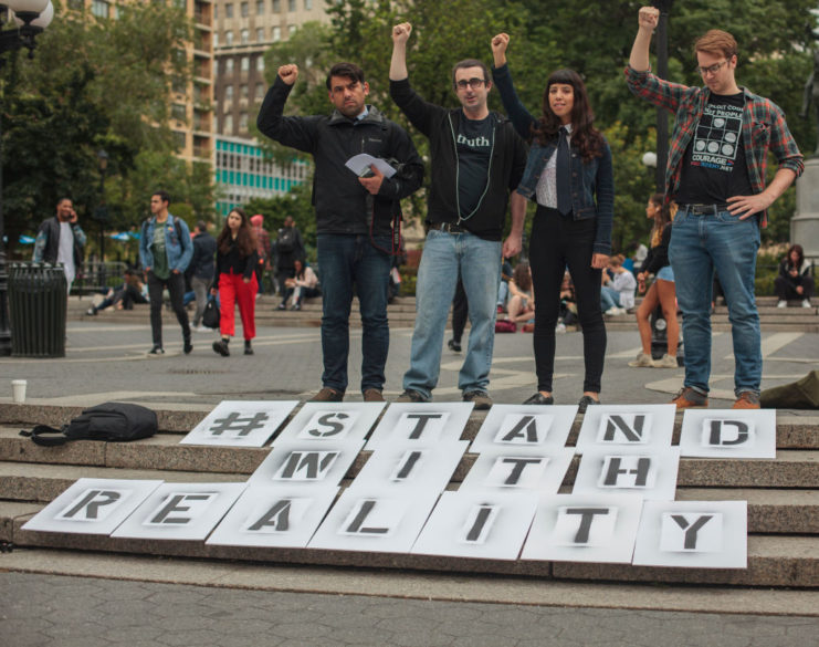 Four individuals standing before a sign that reads "#STANDWITHREALITY"