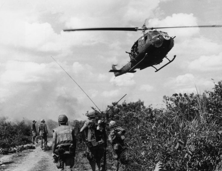 US Marines walking along a path while a US Huey helicopter flies overhead